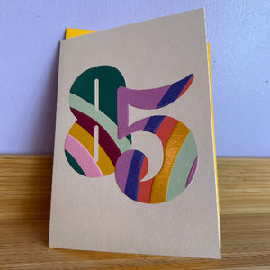 A beige greeting card with a colorful layered paper cutout of the number 85.