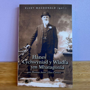 A book titled 'Hanes Cychwyniad y Wladfa ym Mhatagonia' by Elvey MacDonald (gol.) lies on a wooden surface against a light purple wall.