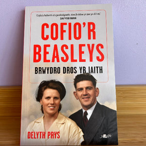A paperback book titled 'Cofio'r Beasleys' by Delyth Prys, featuring a white cover with bold red and black text and a vintage-style portrait of a couple. The book is photographed standing on a wooden surface against a light purple wall.