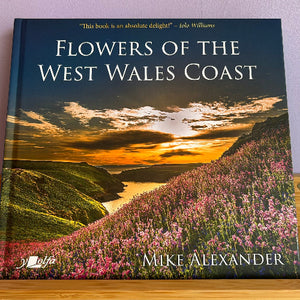 A hardcover book titled "Flowers of the West Wales Coast" by Mike Alexander, featuring a scenic cover photo of a coastal landscape with pink wildflowers, green hills, and a sunset over water. The book is photographed standing upright on a wooden surface against a light wall.