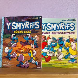 Two Smurf books displayed side by side on a wooden surface against a lavender wall. The left book is titled 'Y Smyrffs Gorau Glas' and the right is 'Y Smyrffs Croeso i Bentr'r Smyrffs'.