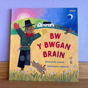 A children's book titled 'BW Y BWGAN BRAIN' by Rhianedd Jewell and Valériane Leblond, featuring a colorful illustration of a scarecrow, placed on a wooden surface against a light purple wall.