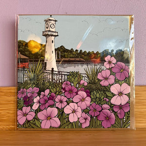 A square card featuring an illustrated lighthouse, water, trees, and a foreground of pink and purple flowers, photographed standing on a wooden surface against a lavender wall.