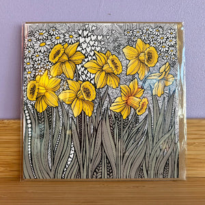 A square floral card featuring yellow daffodils and white daisies with black linework on a gray patterned background, photographed standing upright on a wooden surface against a lavender wall.