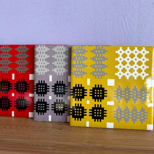 Three square decorative coasters with geometric patterns, shown standing upright on a wooden surface against a light purple wall. The left panel is red with gray and black motifs, the middle panel is gray with white and black motifs, and the right panel is yellow with gray and white motifs.
