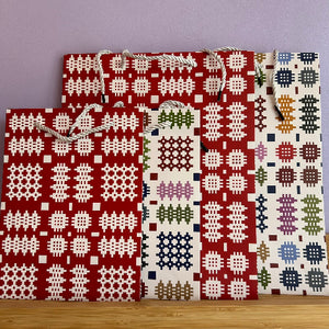 Four woven paper gift bags with rope handles, featuring traditional geometric patterns in red, white, green, blue, and purple, arranged on a wooden surface against a lavender wall.