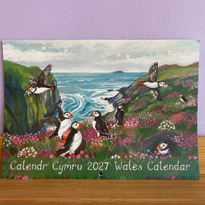 A 2027 Welsh calendar titled 'Calendr Cymru 2027 Wales Calendar' featuring a painted coastal scene with puffins, cliffs, blue sea, and pink flowers, displayed on a wooden surface against a lavender wall.