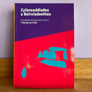A book or booklet standing upright on a wooden surface against a light wall. The cover is split diagonally with a dark purple top section and a bright red bottom section, with teal abstract shapes on the lower half and white text at the top in Welsh.