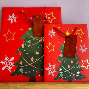 Two red Christmas-themed gift bags with printed gold stars, white snowflakes, and a green decorated Christmas tree, each with a hanging dark red tag.