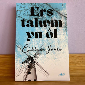 The book cover of 'Ers talwm yn ôl' by Eiddwen Jones, featuring a light blue and white map background with a black windmill illustration and bold black text, photographed standing on a wooden surface against a light purple wall.