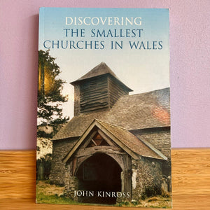 The book 'Discovering the Smallest Churches in Wales' by John Kinross, standing upright on a wooden surface against a light purple wall. The cover features a photograph of a historic stone church with a wooden belfry.