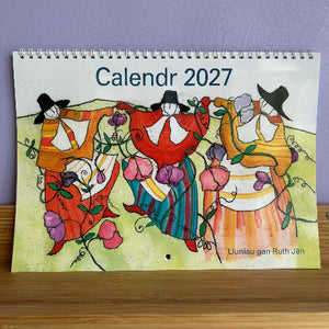 A spiral-bound 2027 calendar displayed on a wooden surface against a lavender wall. The calendar cover features a colorful hand-painted style illustration of three figures in traditional clothing surrounded by flowers, with the text 'Calendr 2027' at the top and 'Liuniau gan Ruth Jún' in the lower right.