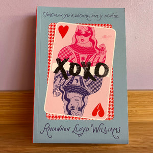 A hardcover book titled 'Torcalon yw'r dechre, dim y diwedd.' by Rhianon Lloyd Williams, featuring a light blue cover with a pink playing card illustration and the word 'XOXO' in black across the center, photographed standing on a wooden surface against a pale pink wall.