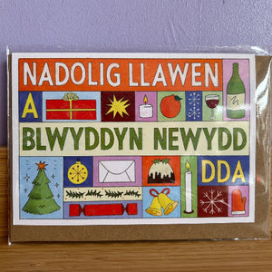 A Christmas greeting card in clear plastic packaging, placed on a wooden surface against a lavender wall. The card features Welsh greetings 'NADOLIG LLAWEN' and 'BLWYDDYN NEWYDD' in large white and green text over a colorful grid of festive illustrations including a Christmas tree, gift box, candle, wine glass, and bells.