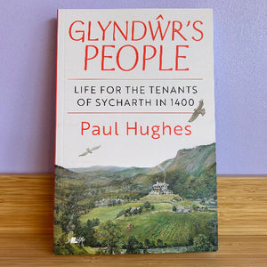 The book 'Glyndwr's People' by Paul Hughes is shown standing upright on a wooden surface against a light purple wall. The cover features a white upper section with red and black text, and a detailed landscape painting of a green valley and castle at the bottom.