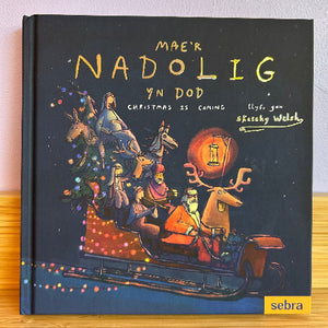A hardcover book titled 'Mae'r Nadolig yn Dod' by sketchy welsh, featuring festive Christmas illustrations on a dark cover, photographed standing upright on a wooden surface against a light wall.