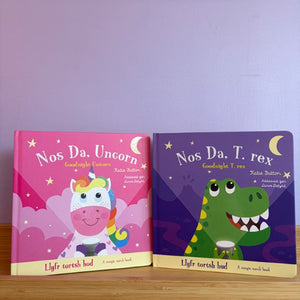 Two children's books titled 'Nos Da, Unicorn' and 'Nos Da, T. rex' on a wooden surface with a purple background.