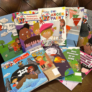 A group of colorful children's books and small booklets fanned out on a brown leather surface. The covers feature playful illustrations of children, animals, and scenes, with Welsh text and bright multicolor artwork.