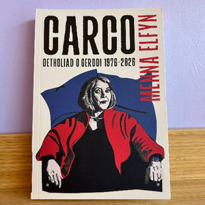 A book titled 'CARCO DETHOLIAD O GERDDI 1976-2026' by Menna Elfyn, featuring a graphic illustration of Menna Elfyn in a red jacket and black top against a blue and cream background, standing on a wooden surface against a light purple wall.