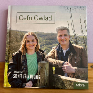 A square hardcover book titled 'Cefn Gwlad' resting on a wooden surface against a light pink wall. The cover features a photograph of a smiling couple outdoors in front of a green landscape, with the text 'GOLYDD SIGNED ERIN HUGHES' and a 'sebra' logo.