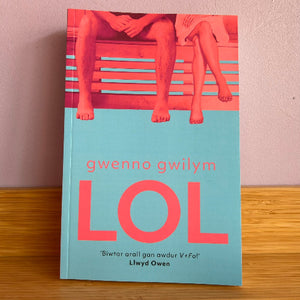 The book cover of 'LOL' by Gwenno Gwilym, featuring a light blue lower half with bold pink lettering and a top section showing the lower legs of two people sitting on a bench, photographed standing on a wooden surface against a wall.