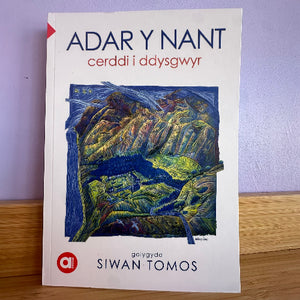 Welsh poetry book titled 'ADAR Y NANT' by Siwan Tomos, featuring a white cover with a colorful mountain landscape illustration and the red 'ai' publisher logo, photographed on a wooden surface against a light purple wall.