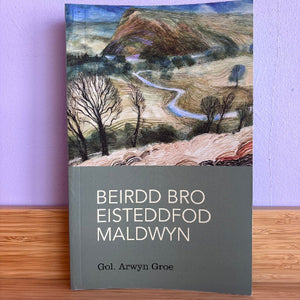 A book titled 'BEIRDD BRO EISTEDDFOD MALDWYN' by Gol. Arwyn Groe, featuring a landscape illustration on the front cover, standing upright on a wooden surface against a light purple wall.