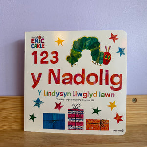 A children's board book titled '123 y Nadolig' by Eric Carle, featuring the iconic green caterpillar and colorful star illustrations on a cream cover, photographed standing on a wooden surface against a light purple wall.