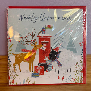 A square Christmas greeting card in its clear plastic shrink-wrap, resting on a wooden surface against a purple wall. The card features a festive winter illustration with the Welsh text 'Nadolig Llawen o bell' at the top.