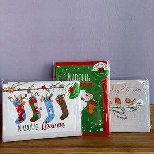 Three Christmas greeting cards in clear plastic packaging, displayed on a wooden surface against a light purple wall. The left card is white with colorful stockings and the text 'NADOLIG Llawen'; the center card is green with a stocking illustration and 'NADOLIG' text; the right card is white with two birds and 'Nadolig Llawen' script.