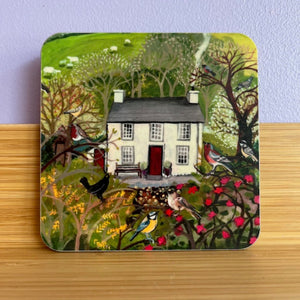Coaster with a design of a house surrounded by trees and birds on a wooden surface.