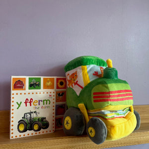 Plush toy train and children's book titled 'Y fferm the farm' on a wooden surface with a purple wall background.