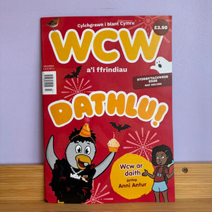 A Welsh language magazine titled 'WCW Dathlu!' featuring a red cover with cartoon illustrations, photographed standing upright on a wooden surface against a light purple wall.
