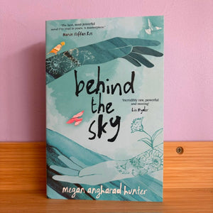The book cover of 'behind the sky' by Megan Anghadad Hunter, featuring a teal and light blue watercolor background with illustrations of hands, birds, and flowers, displayed on a wooden surface against a pink wall.