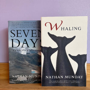 Two paperback books standing upright on a wooden surface against a lavender wall: the front book is 'Whaling' by Nathan Munday with a cream cover and large black whale silhouettes, and the back book is 'Seven Days' by the same author with a dark landscape photo and large black title text.