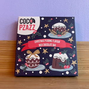 Coco Pzazz Christmas pudding milk chocolate bar packaging on a wooden surface with a purple background