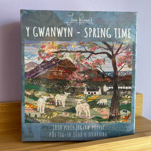 Jigsaw puzzle box titled 'Y Gwanwyn - Spring Time' by Josie Russell on a wooden surface.