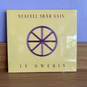 CD cover with a purple symbol on a beige background, against a blue wall.