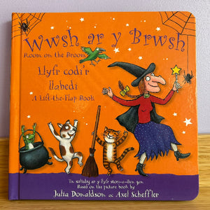 Children's book titled 'Wwsh ar y Brwsh' with illustrations of a witch, cat, and frog on an orange cover.
