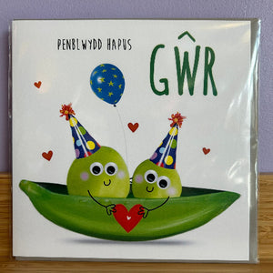 Card with two peas in a pod holding a heart, wearing party hats, and a blue balloon. Text 'Penblwydd Hapus Gwr' on a white background.