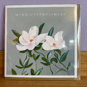 Floral card with white flowers and green leaves on a gray background, featuring the text 'MEWN CYDYMDEIMLAD'.