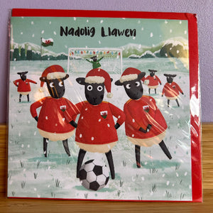 Christmas card with sheep playing football in red jerseys on a snowy field.