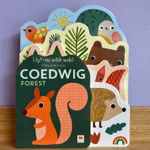 Children's book titled 'Coedwig Forest' with animal illustrations on a wooden surface and purple background.