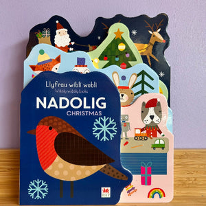 Children's book titled 'Nadolig Christmas' with festive illustrations on a wooden surface.
