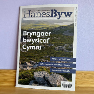 Magazine cover titled 'Hanes Byw' with a landscape photo on a wooden surface and purple wall background