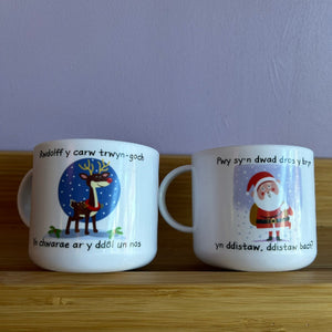 Two mugs with Christmas-themed designs on a wooden surface against a purple wall.