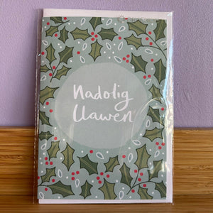 Greeting card with holly design and 'Nadolig Llawen' text on a purple wall background