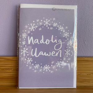 Purple greeting card with 'Nadolig Llawen' text and snowflakes on a wooden surface and purple wall background.