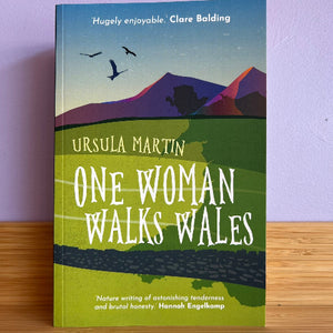 Book cover of 'One Woman Walks Wales' by Ursula Martin on a wooden surface with a purple wall background.