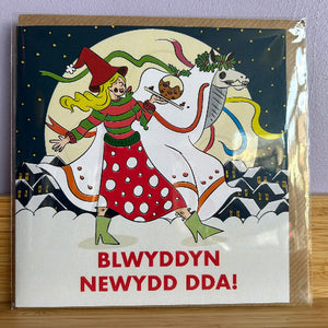 Card with a festive design of a woman in a red dress and green hat, holding a dragon and a pudding, with text 'BLWYDDYN NEWYDD DDA!' on a snowy landscape.